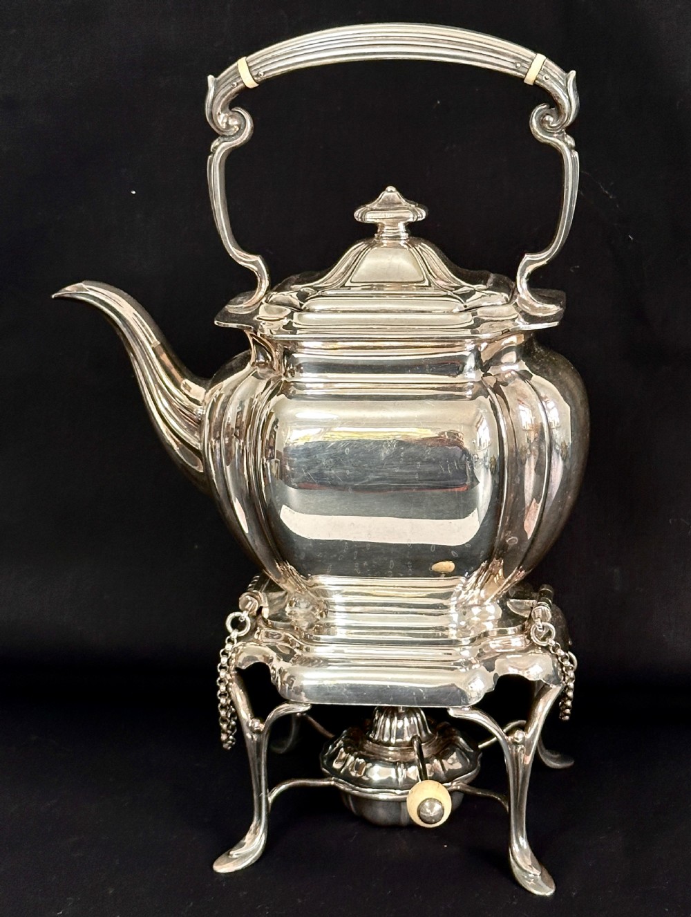 A silver-plated tea kettle with an overhead handle and spirit burner resting on a matching carved four-legged stand.