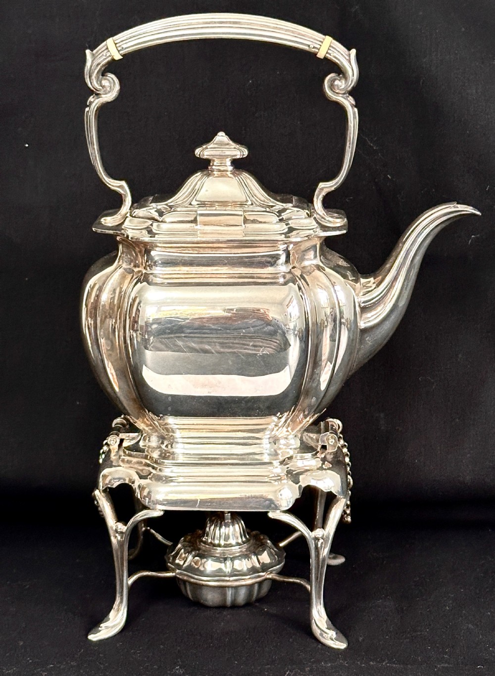 A silver-plated tea kettle with an overhead handle and spirit burner resting on a matching carved four-legged stand. View III