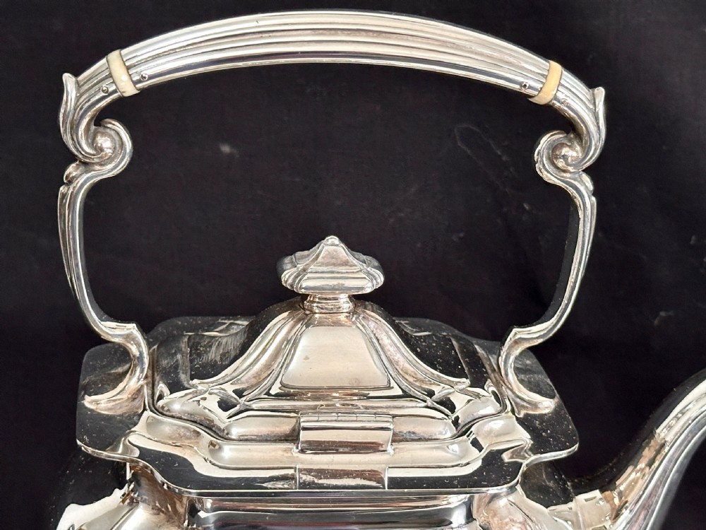 A silver-plated tea kettle with an overhead handle and spirit burner resting on a matching carved four-legged stand. View V