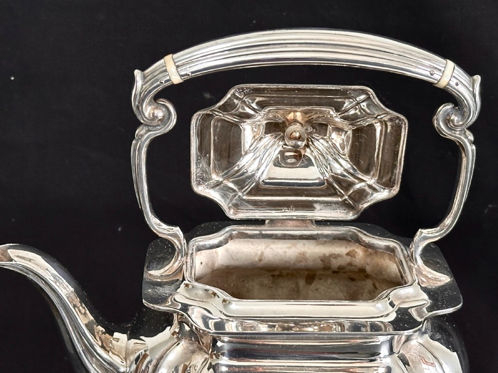 A silver-plated tea kettle with an overhead handle and spirit burner resting on a matching carved four-legged stand. View VI