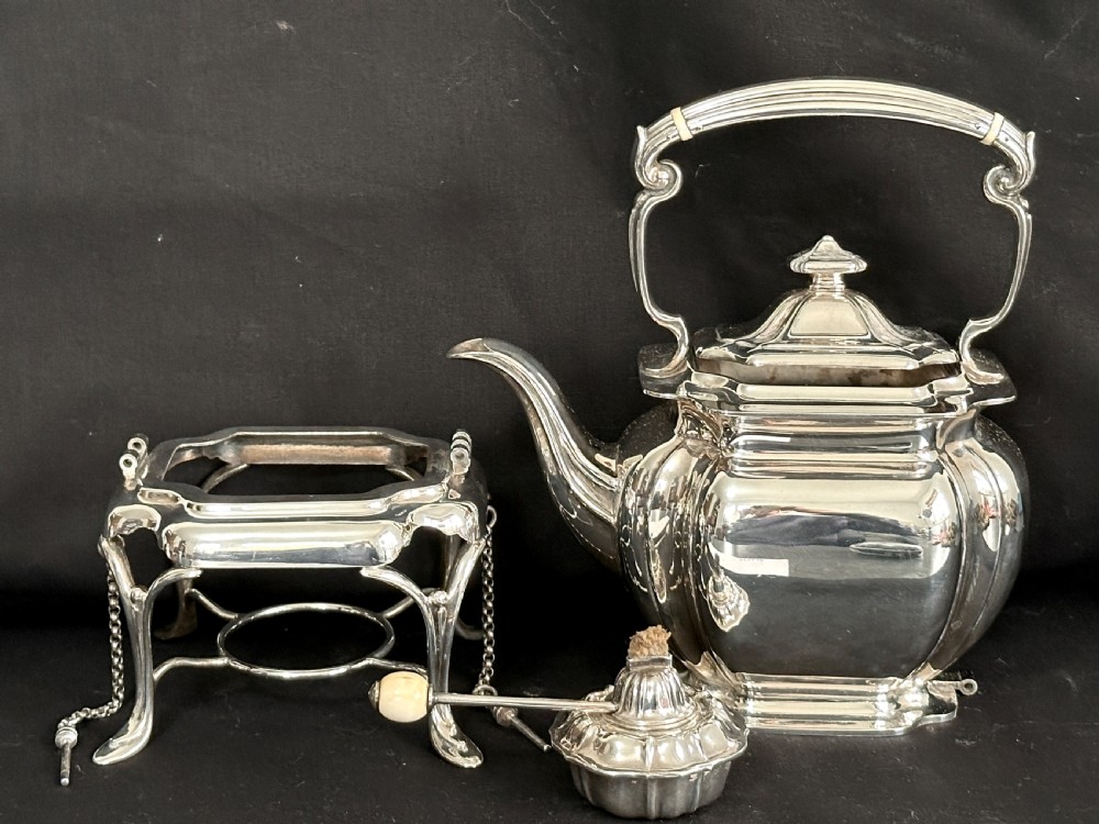 A silver-plated tea kettle with an overhead handle and spirit burner resting on a matching carved four-legged stand. View VII