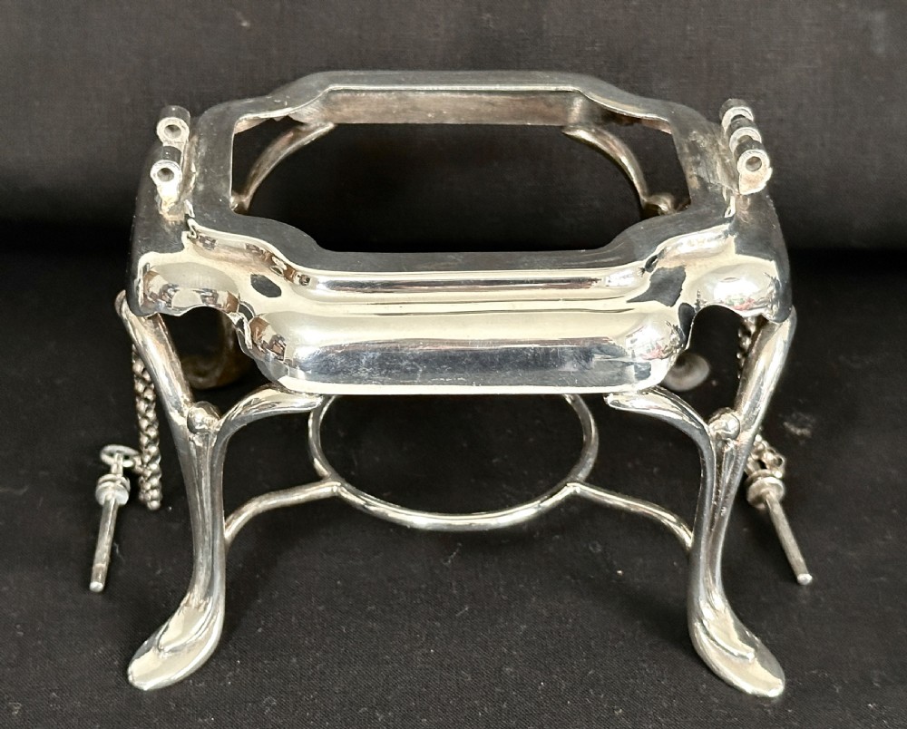 A silver-plated tea kettle with an overhead handle and spirit burner resting on a matching carved four-legged stand. View VIII