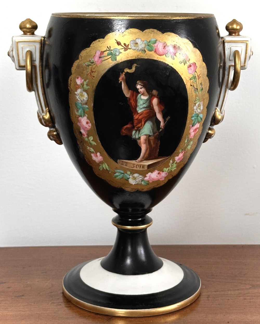 A black French porcelain goblet vase featuring gilt handles, a flared pedestal base, and a central painted medallion depicting a classical figure labelled Le Soir.