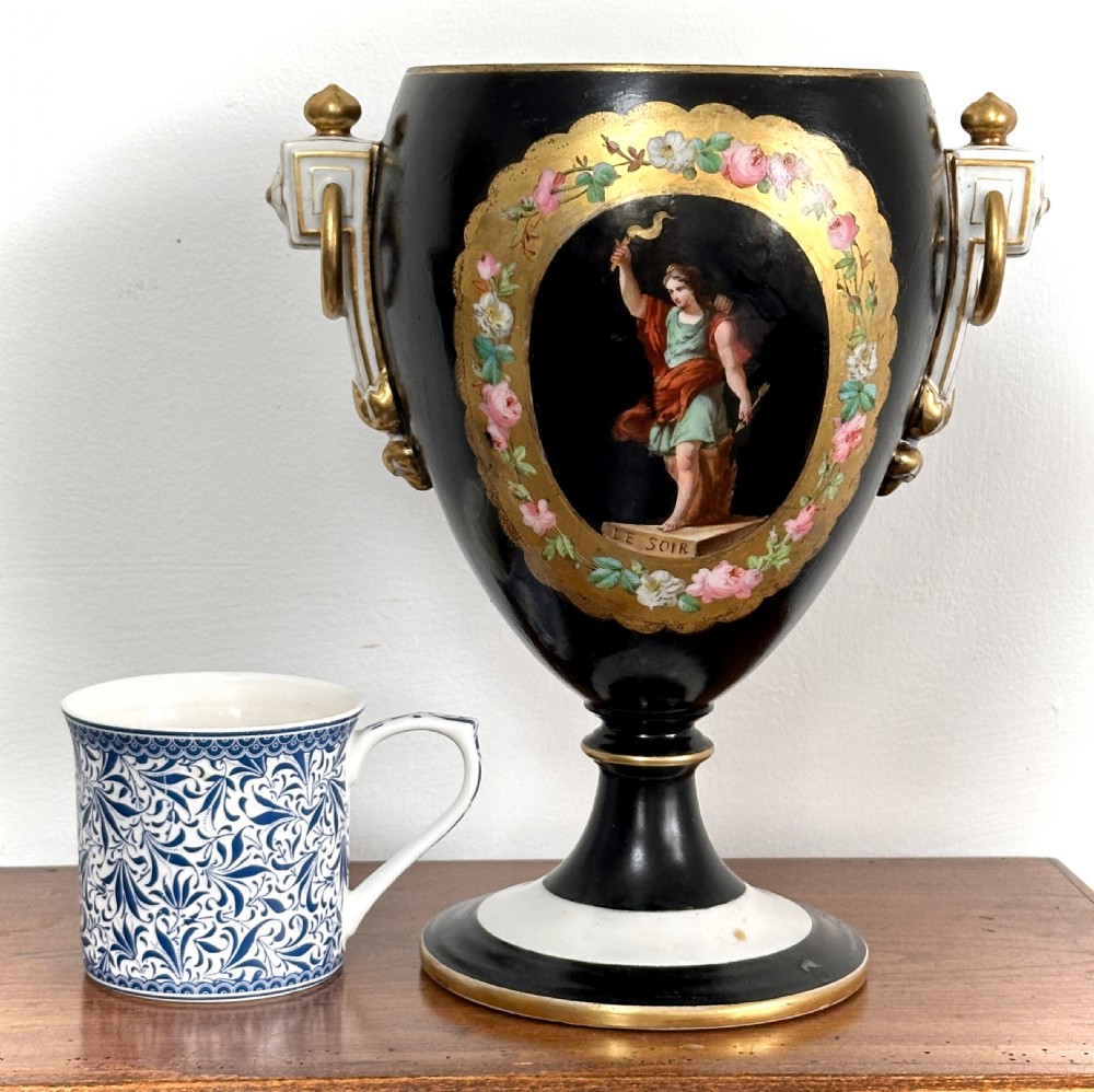 A black French porcelain goblet vase featuring gilt handles, a flared pedestal base, and a central painted medallion depicting a classical figure labelled Le Soir. View IX