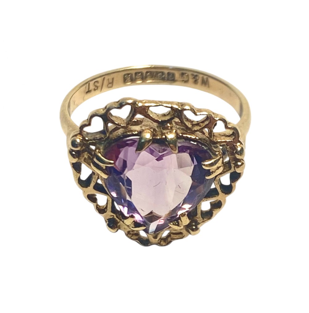 A yellow gold ring featuring a heart-cut purple amethyst set within an openwork heart-motif border and a hallmarked band.