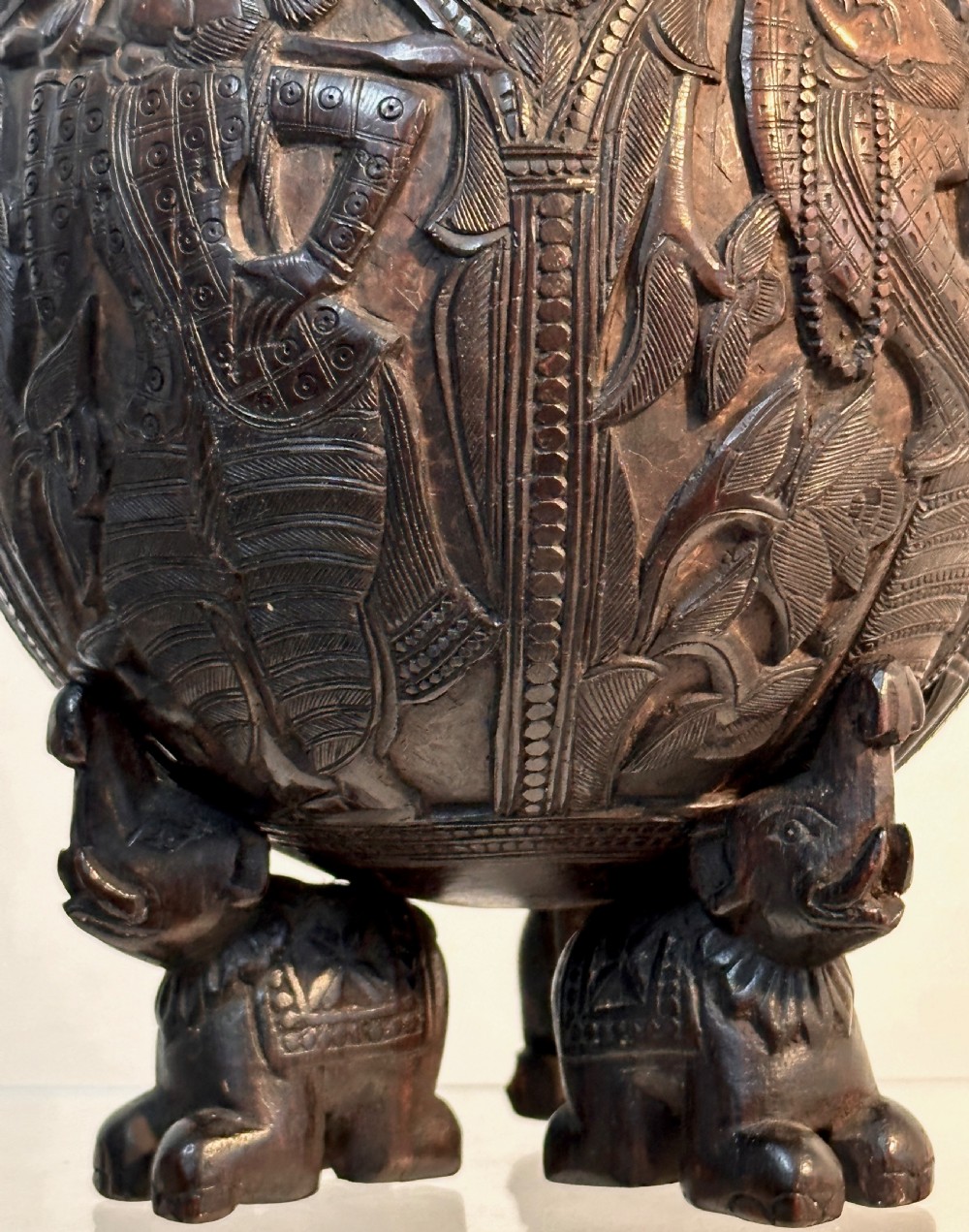 An intricately carved dark brown coconut shell vessel featuring relief figures and a bird-shaped lid handle, resting on carved legs. View II