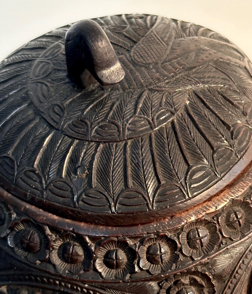 An intricately carved dark brown coconut shell vessel featuring relief figures and a bird-shaped lid handle, resting on carved legs. View III
