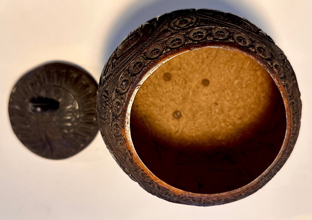 An intricately carved dark brown coconut shell vessel featuring relief figures and a bird-shaped lid handle, resting on carved legs. View IV