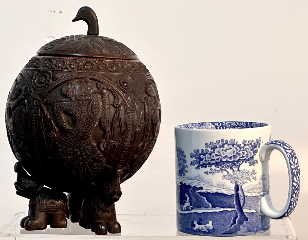 An intricately carved dark brown coconut shell vessel featuring relief figures and a bird-shaped lid handle, resting on carved legs. View VIII