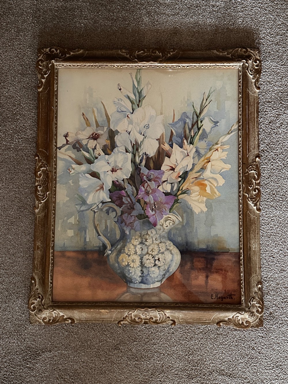 A framed watercolour painting depicting white and purple gladioli in an ornate ceramic jug, presented within a carved gilt frame. View V