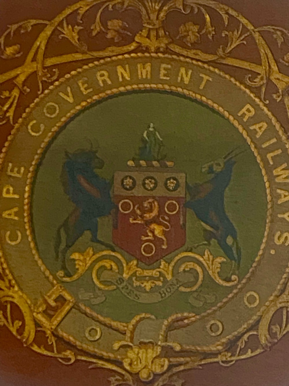 An oval dark wood plaque inscribed with Cape Government Railways featuring a painted coat of arms and gilded floral scrollwork. View IV