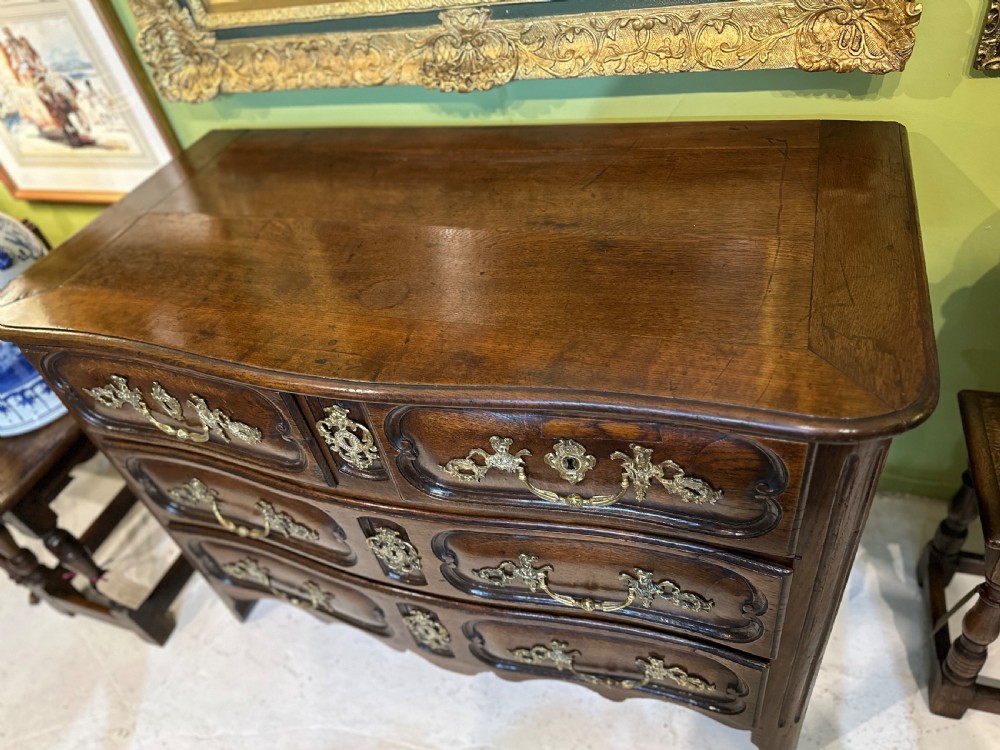 An 18th-century French oak serpentine chest of drawers featuring three rows of moulded drawer fronts and decorative brass handles. View IV