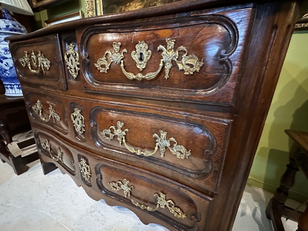 An 18th-century French oak serpentine chest of drawers featuring three rows of moulded drawer fronts and decorative brass handles. View VI