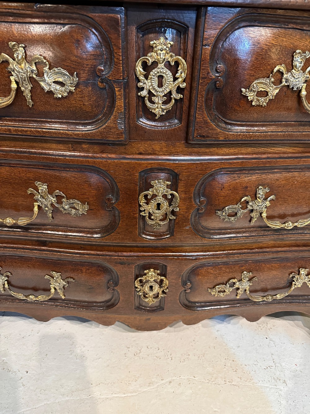 An 18th-century French oak serpentine chest of drawers featuring three rows of moulded drawer fronts and decorative brass handles. View VII