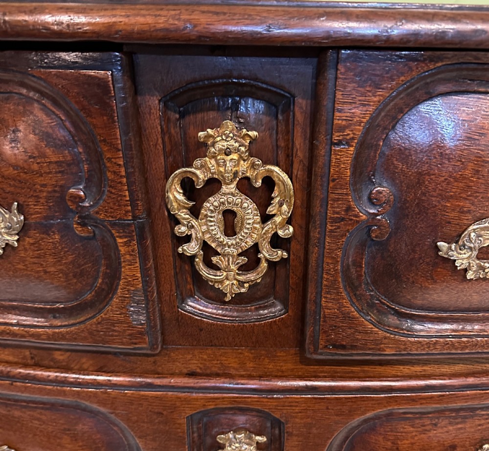An 18th-century French oak serpentine chest of drawers featuring three rows of moulded drawer fronts and decorative brass handles. View XIV
