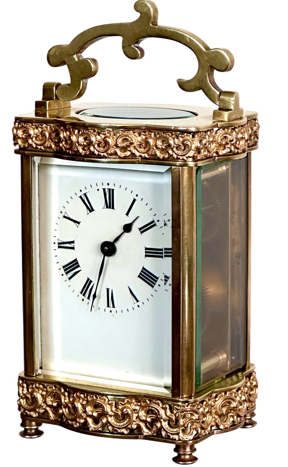 An antique French brass carriage clock featuring serpentine glass panels, scrollwork borders, a white enamel dial with Roman numerals, and a carrying handle.