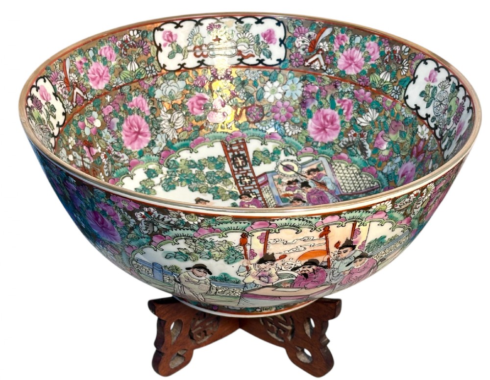 A rounded Chinese Famille Rose porcelain bowl decorated with vibrant pink floral enamelling and figural court scenes, resting on a carved dark wooden stand.