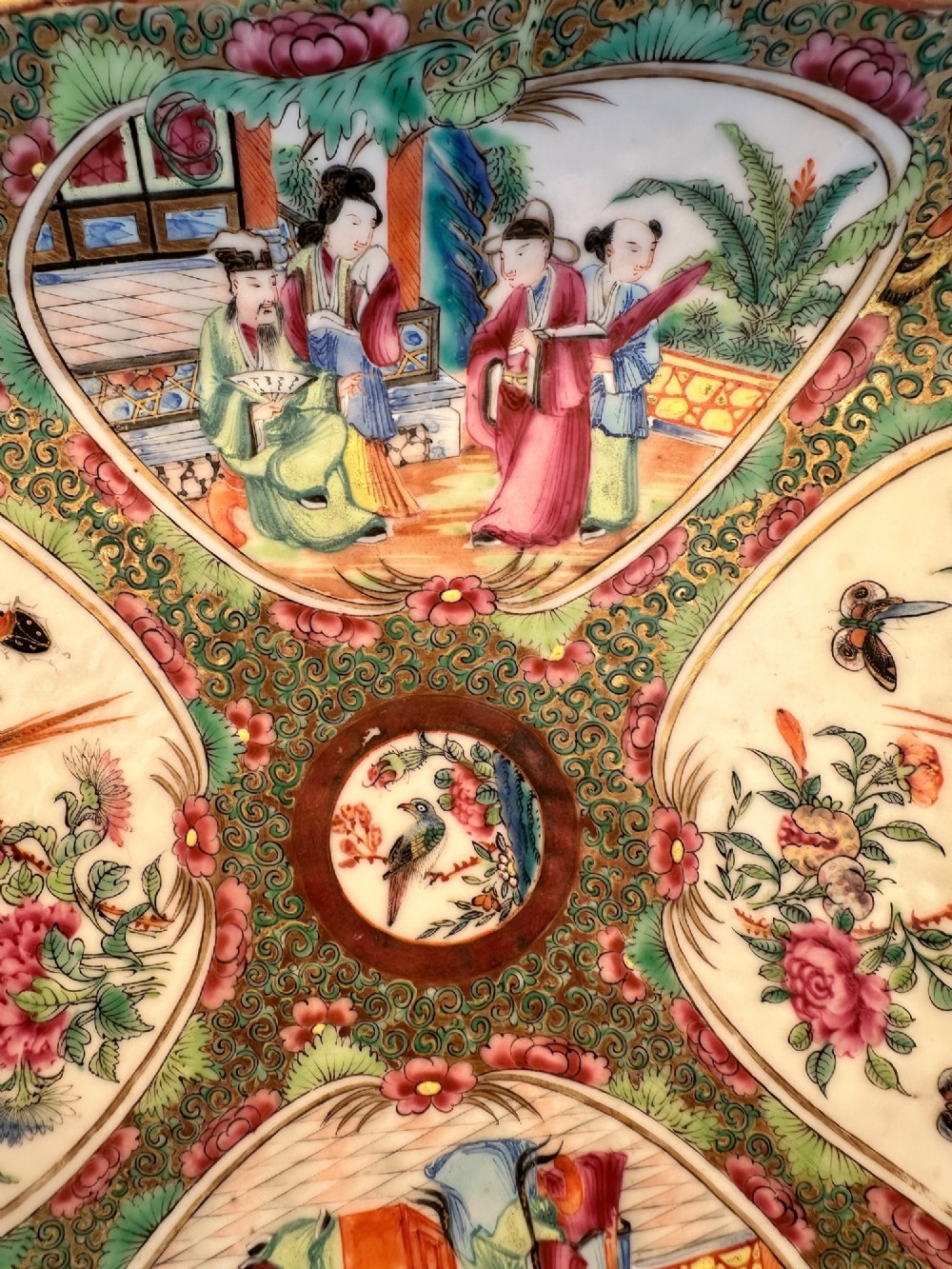 A square Chinese porcelain dish with lobed corners featuring painted reserves of figural court scenes, birds, and flowers set against a dense green scroll and butterfly ground. View II