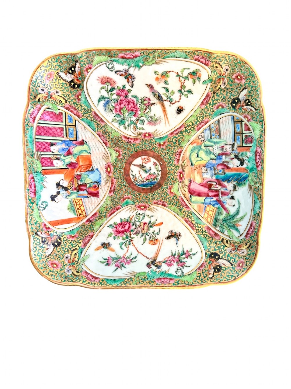 A square Chinese porcelain dish with lobed corners featuring painted reserves of figural court scenes, birds, and flowers set against a dense green scroll and butterfly ground.
