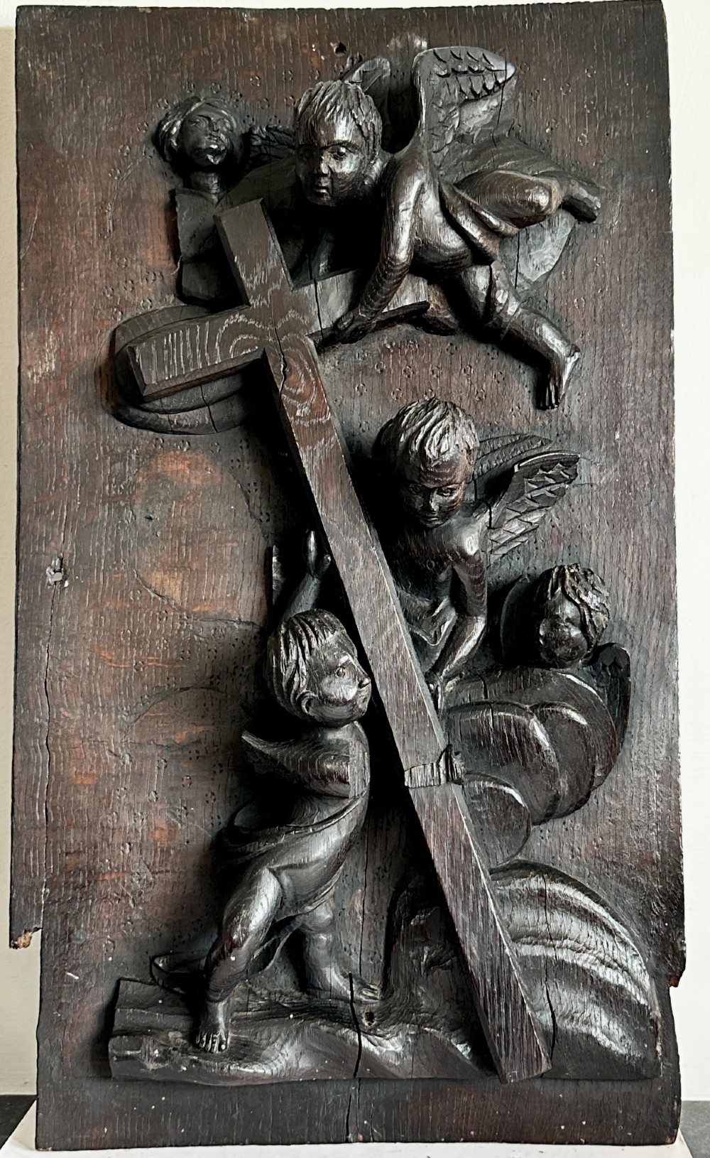 A rectangular dark oak timber panel featuring a high-relief carving of cherubs surrounding a large wooden cross. View III