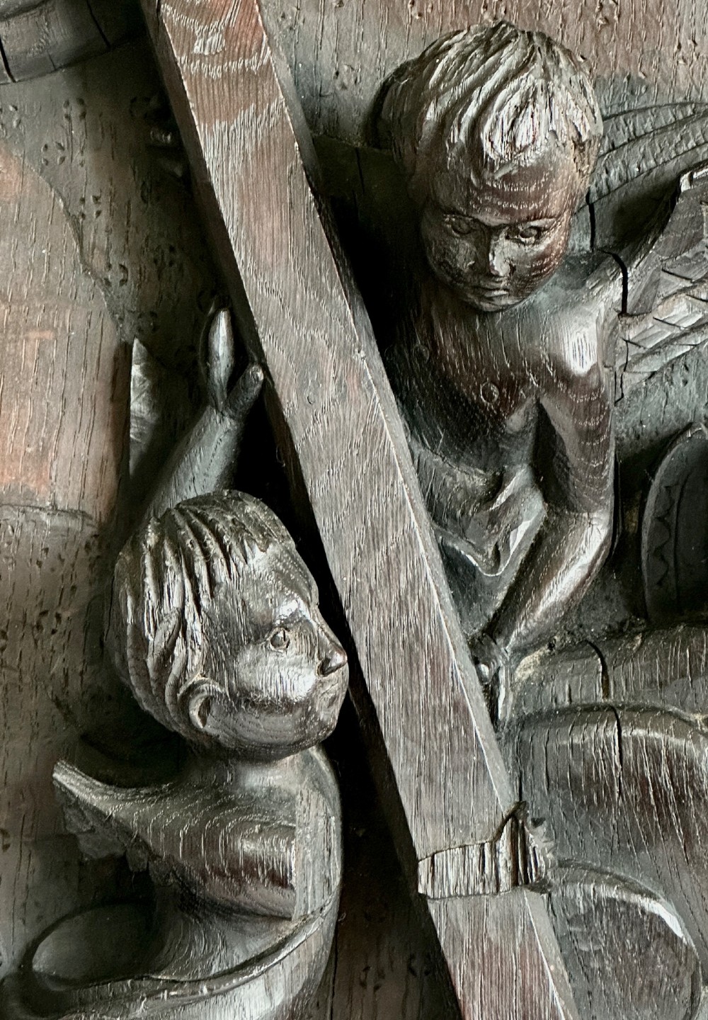 A rectangular dark oak timber panel featuring a high-relief carving of cherubs surrounding a large wooden cross. View IV