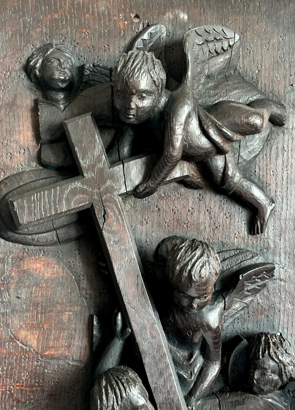 A rectangular dark oak timber panel featuring a high-relief carving of cherubs surrounding a large wooden cross. View V