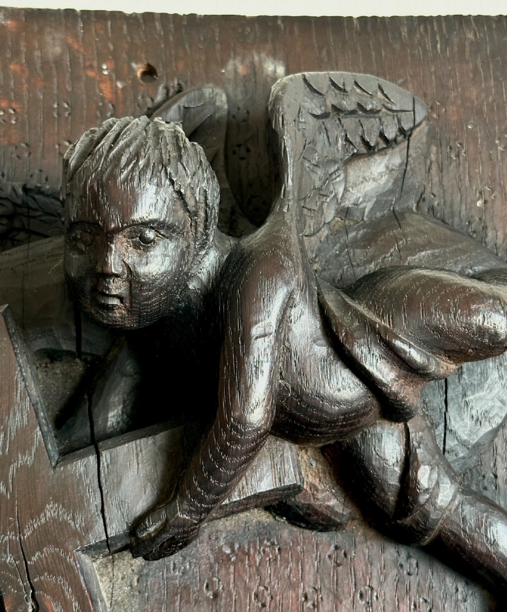 A rectangular dark oak timber panel featuring a high-relief carving of cherubs surrounding a large wooden cross. View X