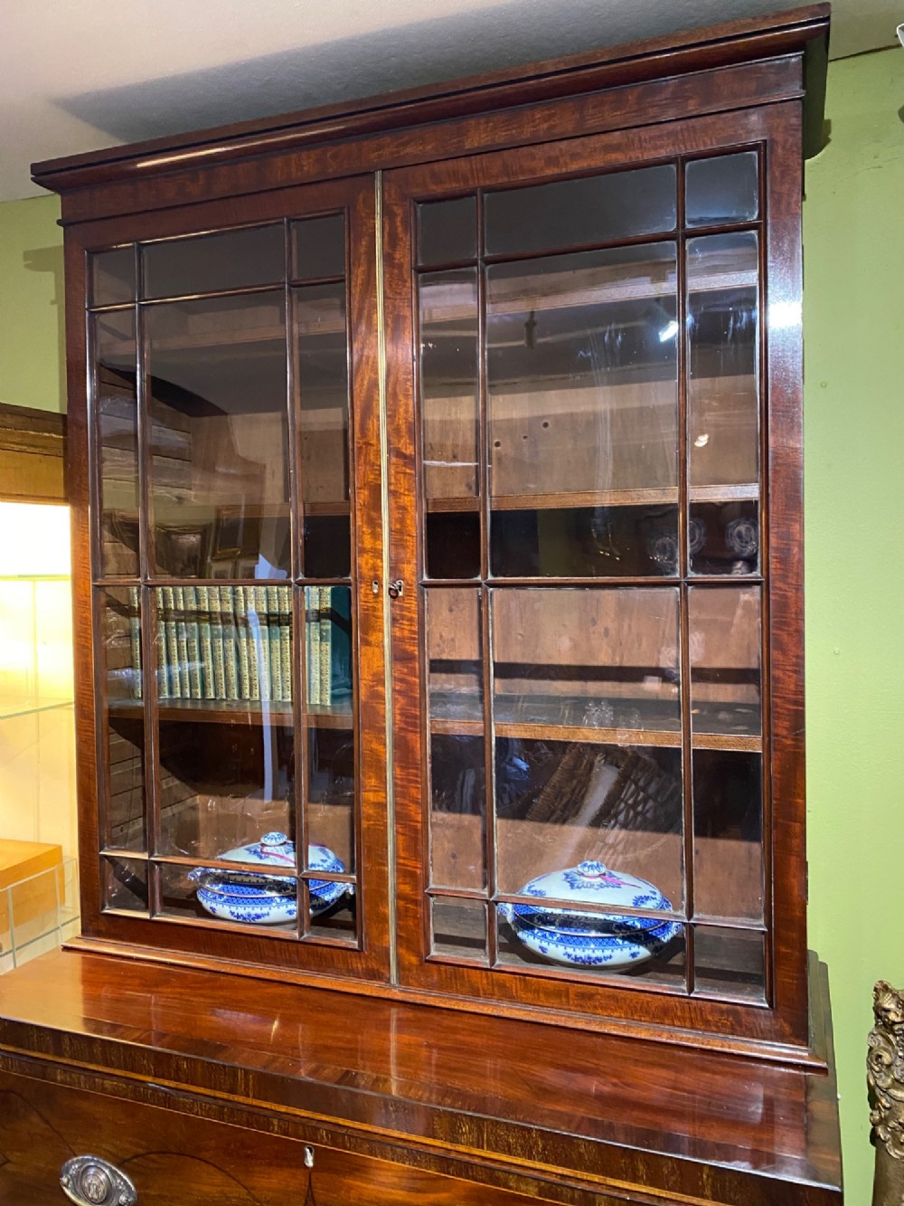An antique Regency mahogany secretaire bookcase featuring an upper two-door glazed cabinet above a lower chest of drawers with decorative inlay and brass oval drop handles. View IX