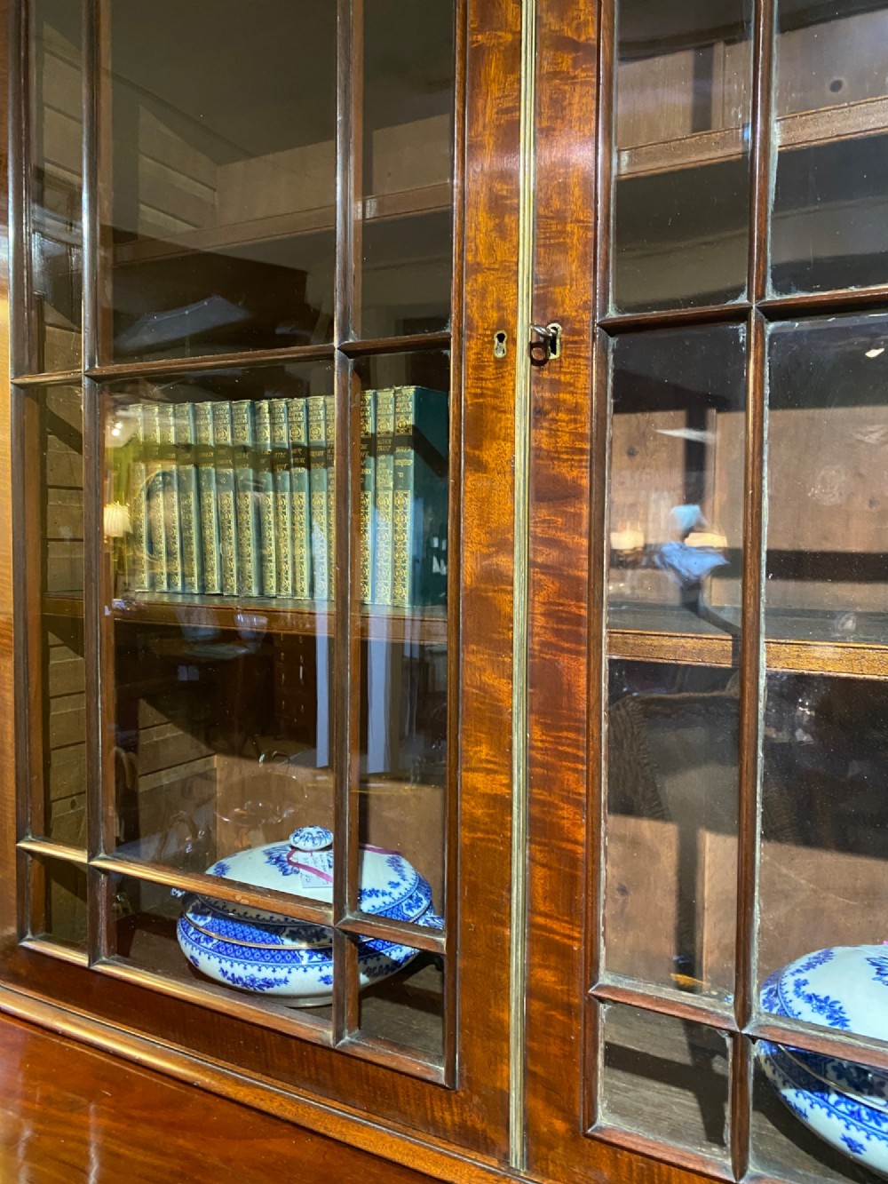 An antique Regency mahogany secretaire bookcase featuring an upper two-door glazed cabinet above a lower chest of drawers with decorative inlay and brass oval drop handles. View X