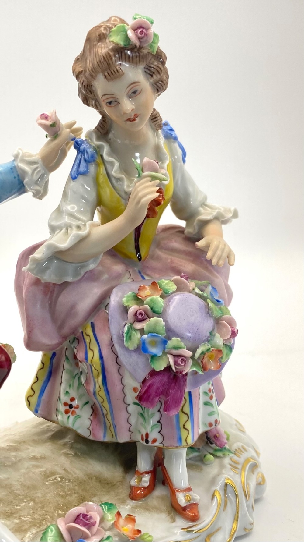 A Sitzendorf porcelain figurine group depicting an 18th-century courtly couple seated together with flower baskets on a scrollwork base with gold detailing. View IV