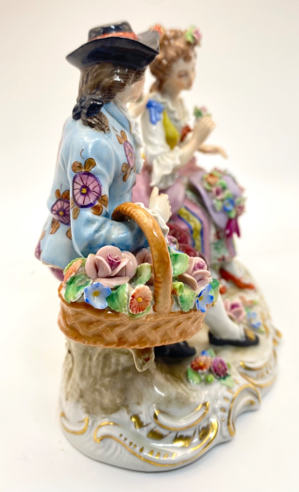 A Sitzendorf porcelain figurine group depicting an 18th-century courtly couple seated together with flower baskets on a scrollwork base with gold detailing. View VI