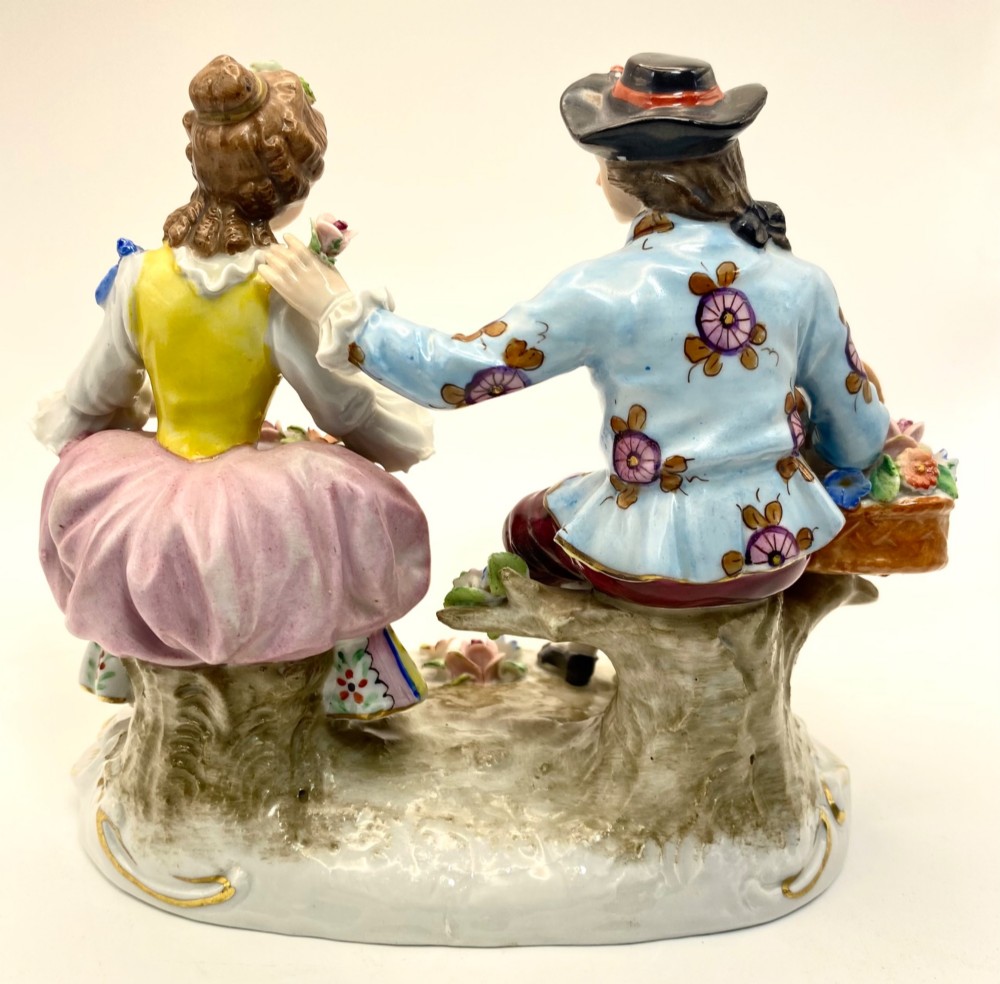 A Sitzendorf porcelain figurine group depicting an 18th-century courtly couple seated together with flower baskets on a scrollwork base with gold detailing. View VII