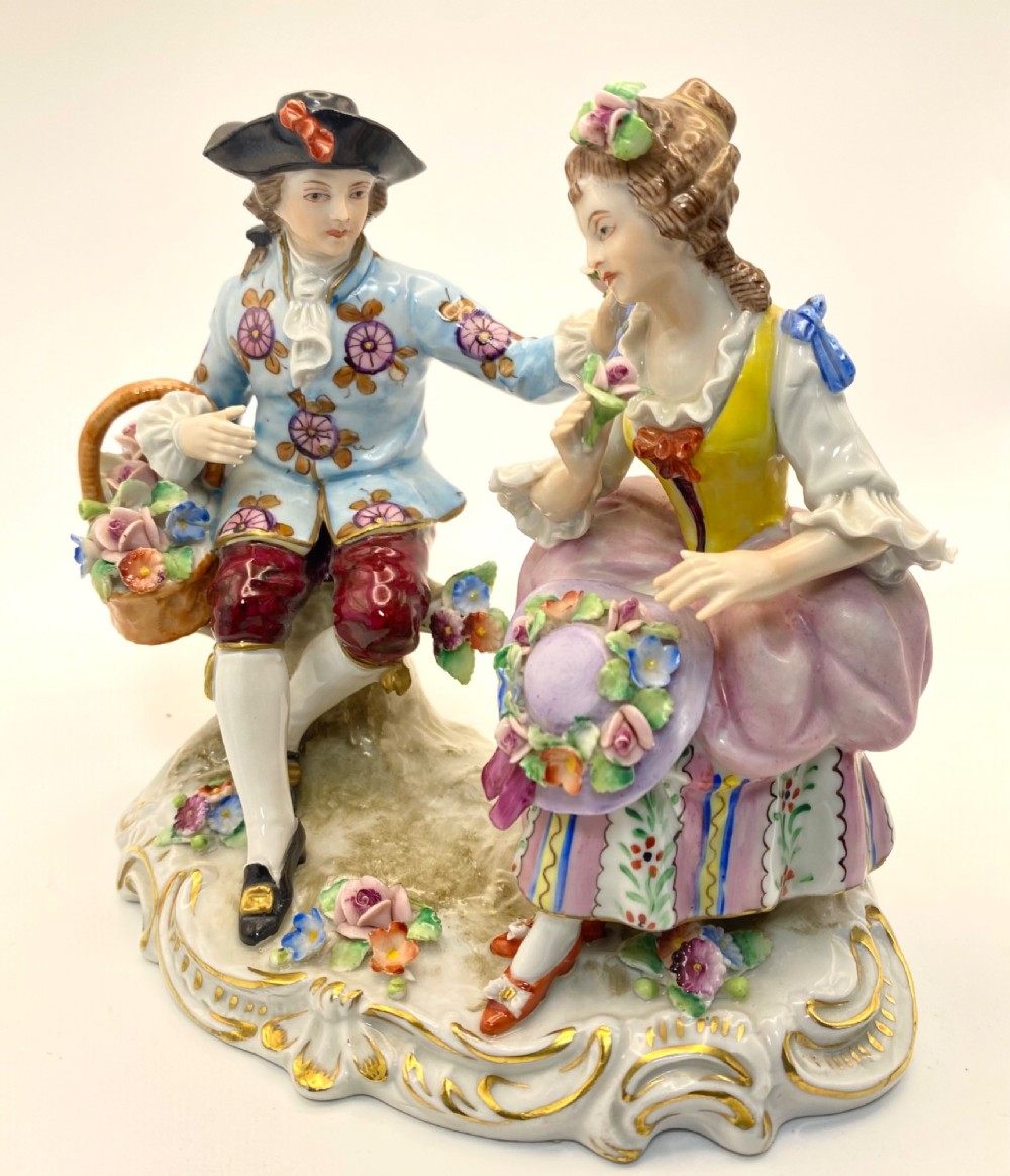 A Sitzendorf porcelain figurine group depicting an 18th-century courtly couple seated together with flower baskets on a scrollwork base with gold detailing.
