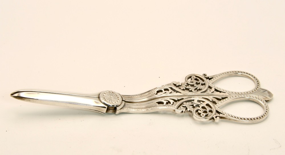 A pair of ornate silver grape scissors featuring pierced scrollwork handles, displayed inside an open blue velvet-lined fitted case. View II
