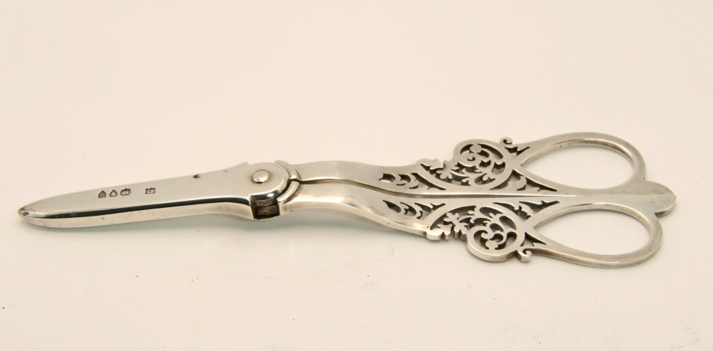 A pair of ornate silver grape scissors featuring pierced scrollwork handles, displayed inside an open blue velvet-lined fitted case. View IV