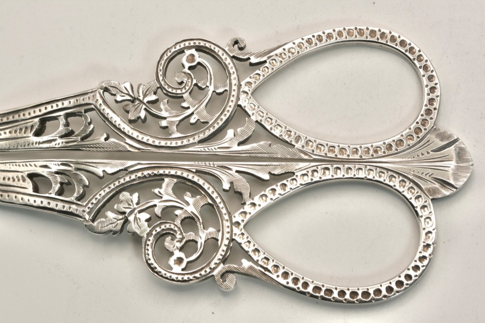 A pair of ornate silver grape scissors featuring pierced scrollwork handles, displayed inside an open blue velvet-lined fitted case. View V