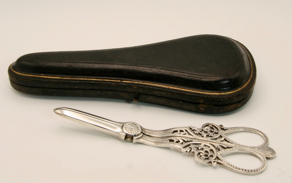 A pair of ornate silver grape scissors featuring pierced scrollwork handles, displayed inside an open blue velvet-lined fitted case. View VIII