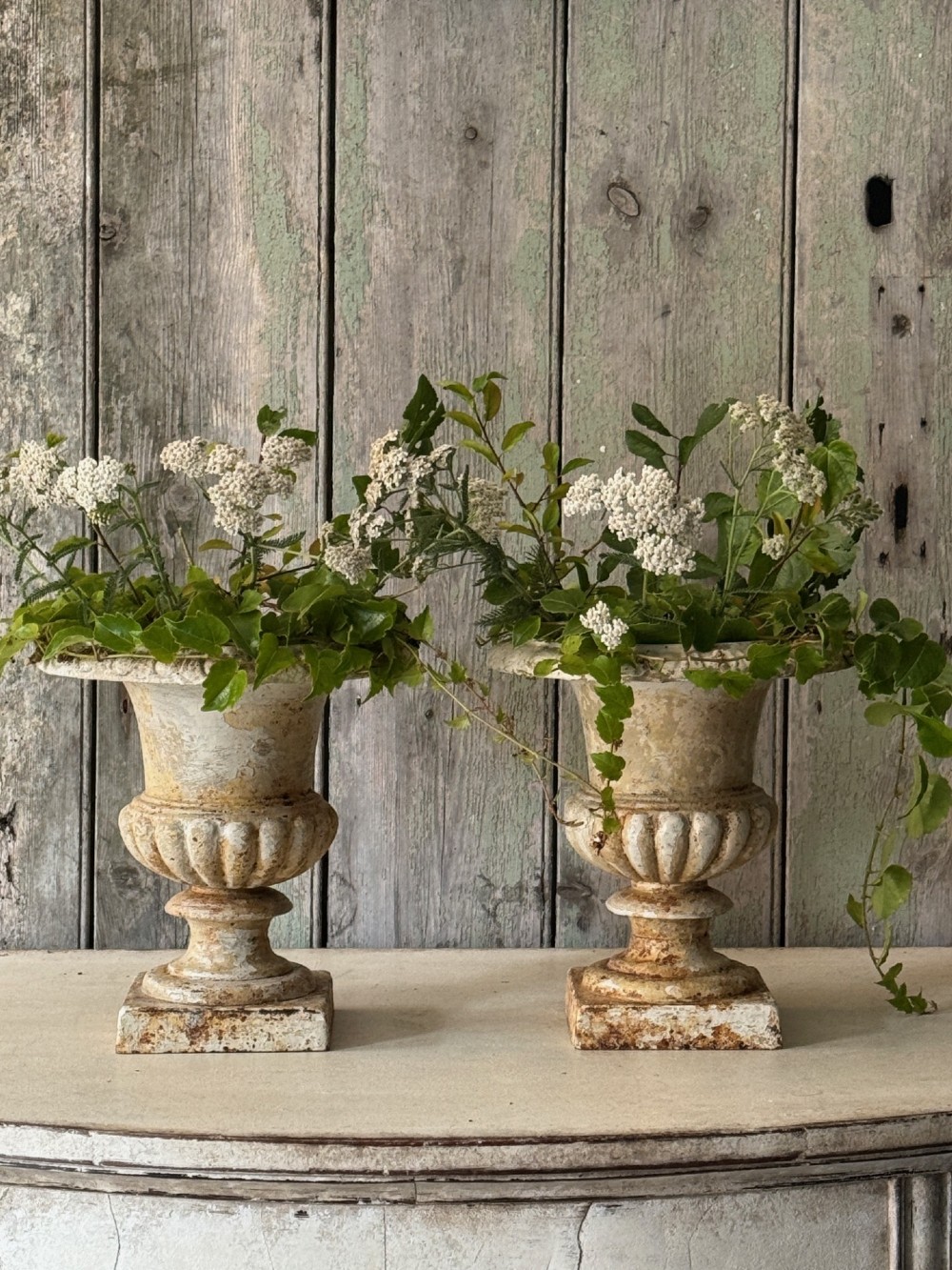 A pair of antique French cast iron garden urns with fluted bodies, square bases, and weathered off-white painted finishes showing natural rust patina.