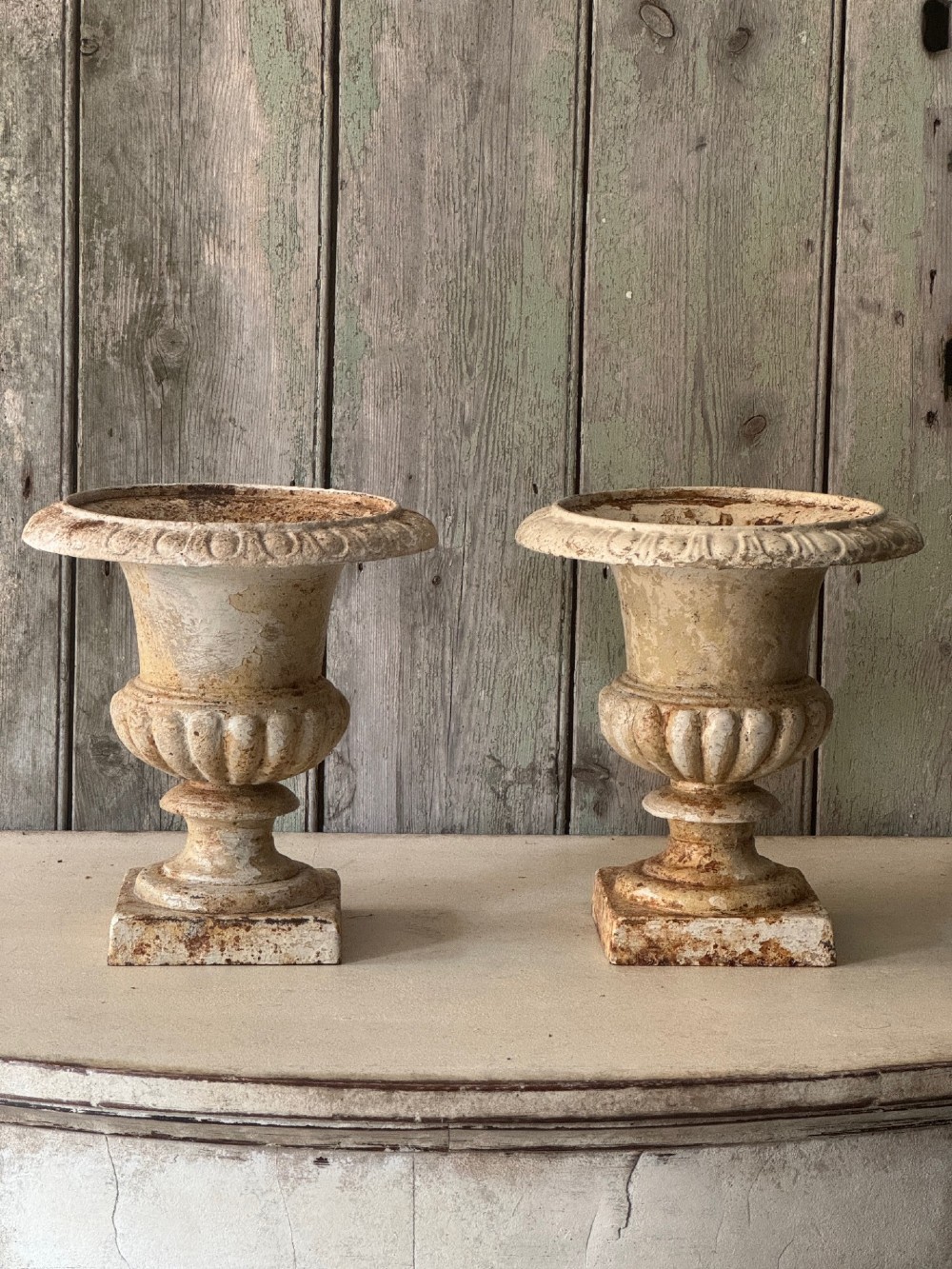 A pair of antique French cast iron garden urns with fluted bodies, square bases, and weathered off-white painted finishes showing natural rust patina. View II