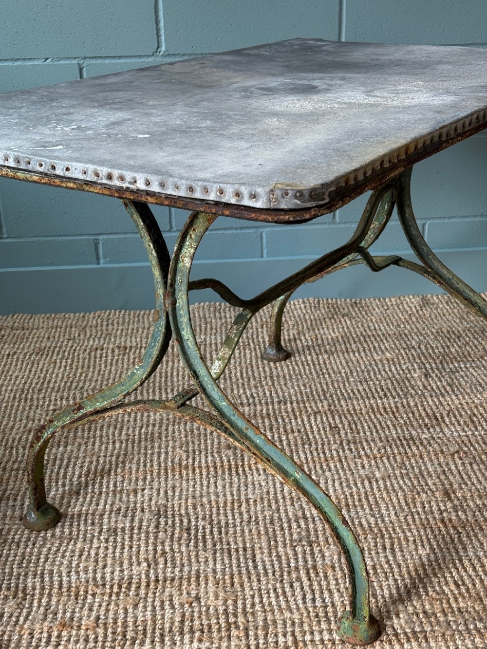 A rectangular French dining table with a riveted zinc top and dark metal frame, surrounded by six black slatted folding bistro chairs on gravel. View II