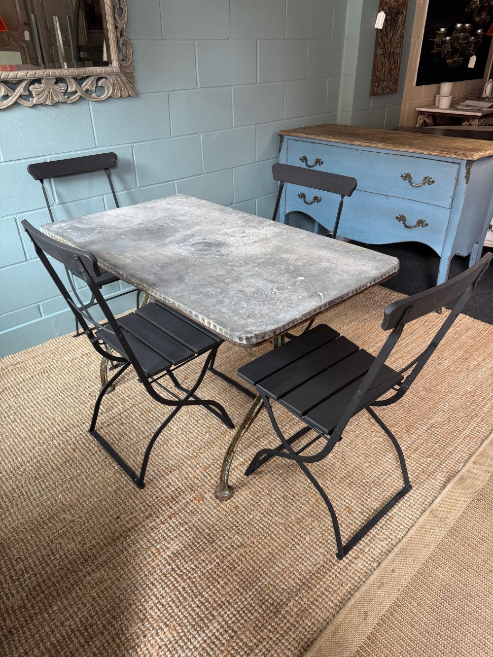 A rectangular French dining table with a riveted zinc top and dark metal frame, surrounded by six black slatted folding bistro chairs on gravel. View V