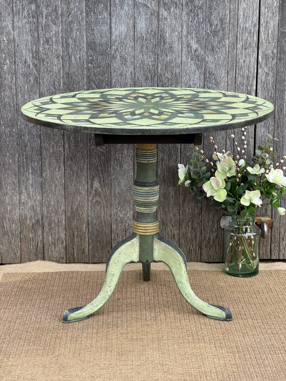 A Victorian circular tilt-top side table featuring hand-painted geometric patterns in sage green, olive, and cream resting on a turned stem with tripod legs.