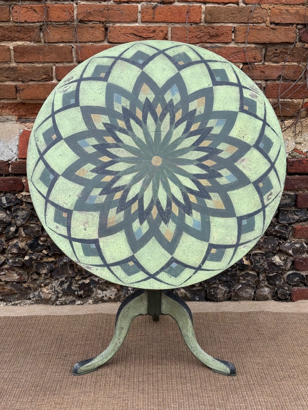 A Victorian circular tilt-top side table featuring hand-painted geometric patterns in sage green, olive, and cream resting on a turned stem with tripod legs. View II