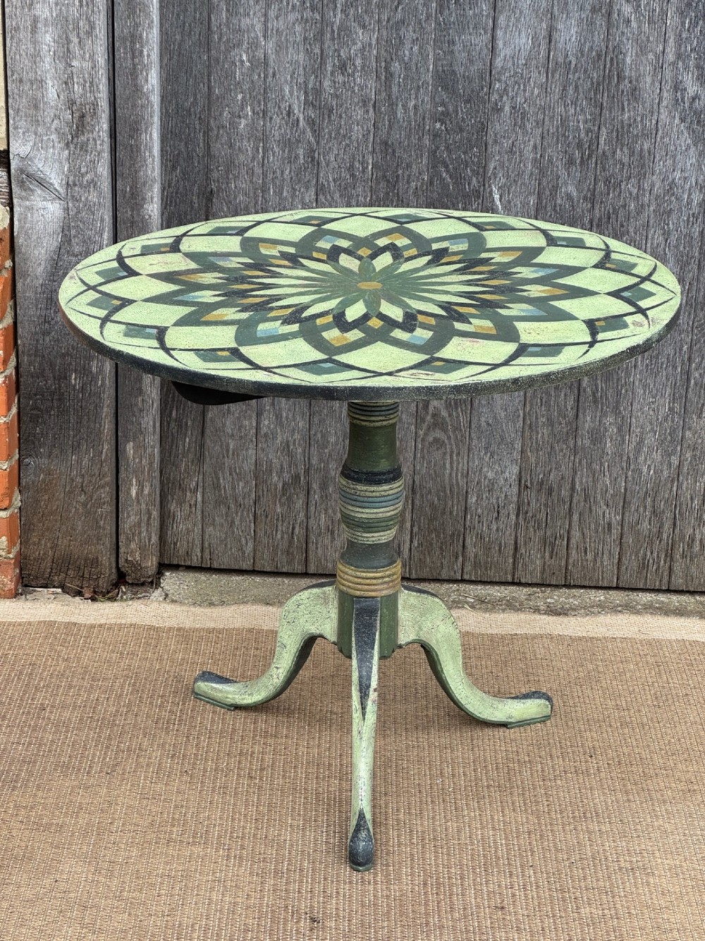 A Victorian circular tilt-top side table featuring hand-painted geometric patterns in sage green, olive, and cream resting on a turned stem with tripod legs. View III