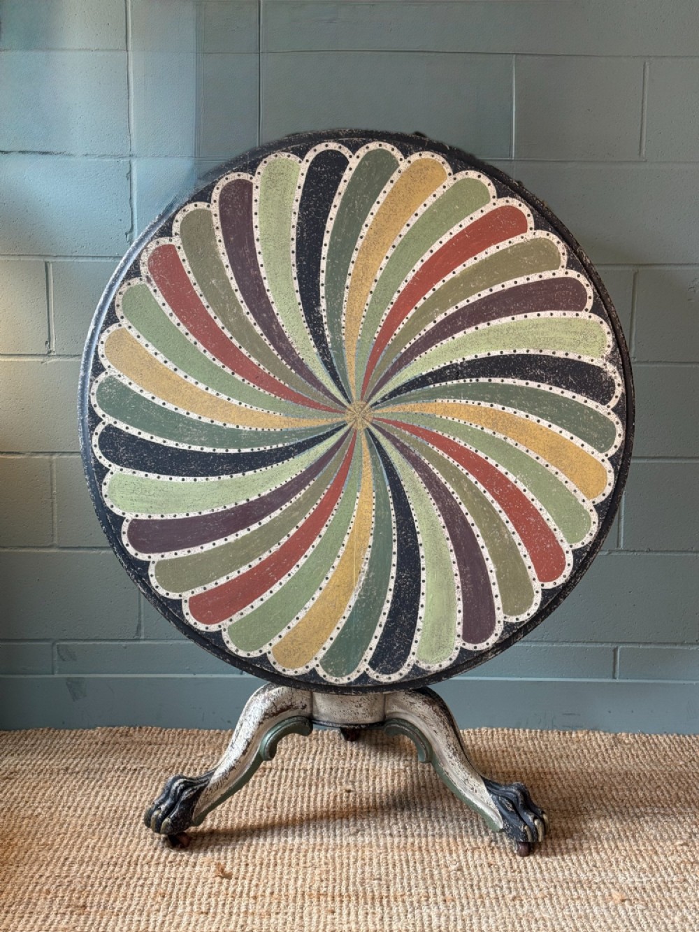 A Victorian circular tilt-top table featuring a hand-painted swirling polychrome petal pattern across its surface, resting on a distressed carved tripod base.