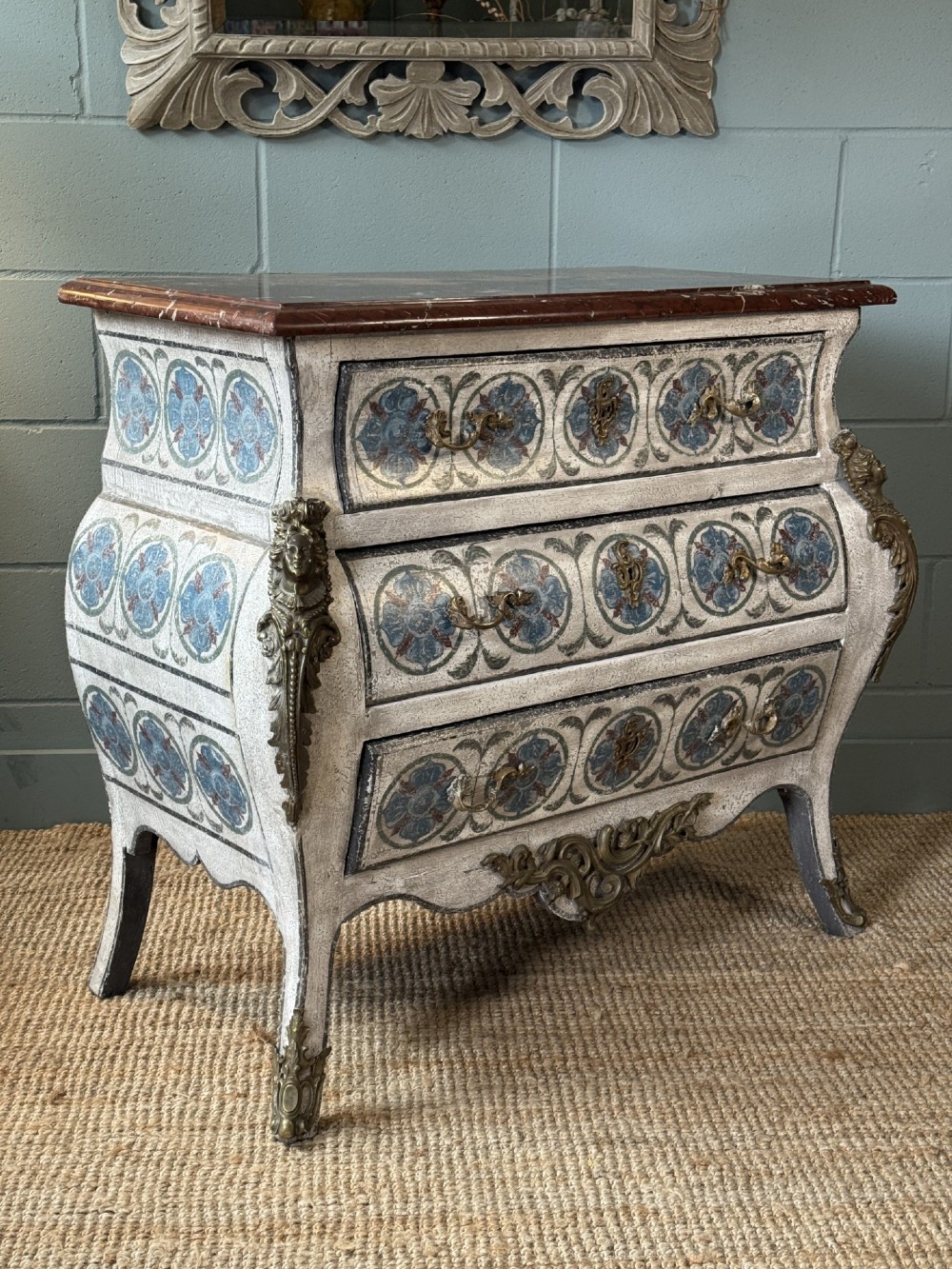 A nineteenth-century French bombee chest of drawers featuring a later hand-painted pattern, a dark marble top, and ornate gilt bronze mounts.
