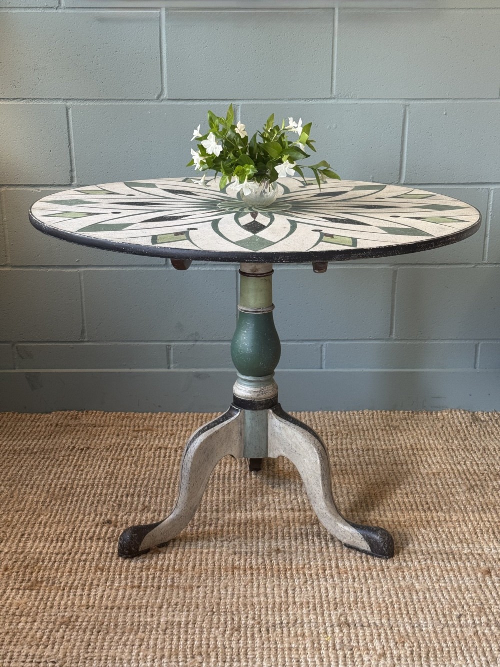 A Victorian circular tilt-top table with a turned tripod pedestal base and hand-painted geometric petal design in green, cream, and black.