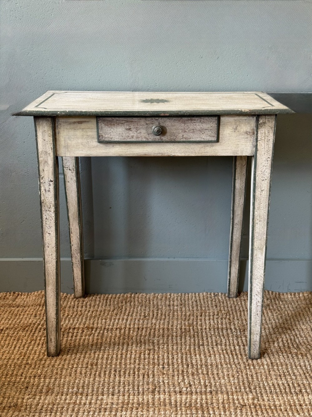 An off-white painted French provincial lamp table with dark trimmed borders, a star motif top, tapered legs, and a single drawer.