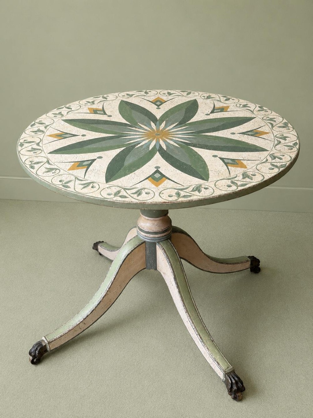 A circular Victorian tilt-top table featuring hand-painted geometric and botanical foliage patterns in green and yellow on a cream ground, resting on a tripod base with brass claw feet. View II