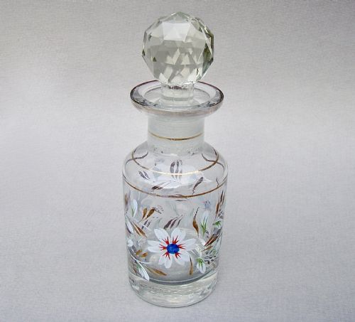 fig 1. A clear glass cologne bottle featuring hand-painted white floral enamel motifs, gilded leaf accents, and a faceted glass stopper.